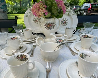 May include: A white tea set with floral designs on the cups and teapot. The set includes a teapot, creamer, sugar bowl, and six cups and saucers. The tea set is on a white tablecloth with a floral pattern.
