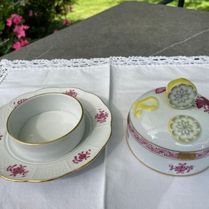 May include: A white porcelain butter dish with a pink floral design. The dish has a lid with a raised design of lemons and a small, round, white porcelain dish inside.
