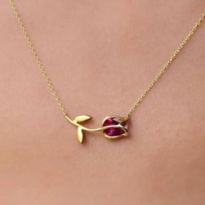 Può includere: Una collana con catena d'oro con un piccolo ciondolo a forma di rosa. La rosa è d'oro e ha una pietra preziosa rossa al centro.