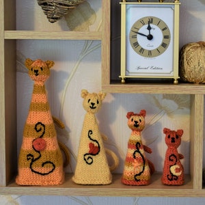 May include: A wooden shelf unit with four shelves displays a variety of items, including a heart-shaped wreath, a clock, a ball of yarn, and four knitted cat figurines. The figurines are in shades of orange, brown, and yellow.