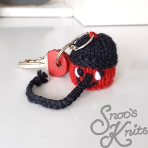 May include: A black and red knitted keychain with a black yarn tail. The keychain features a white face with black eyes and a red nose.