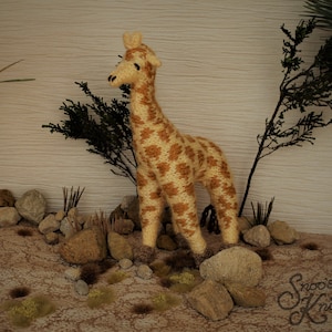May include: A knitted giraffe toy in cream and brown, with a spotted pattern. The giraffe stands on a sandy surface with rocks and small plants, with a backdrop of greenery and a light-coloured wall. The toy is handmade.