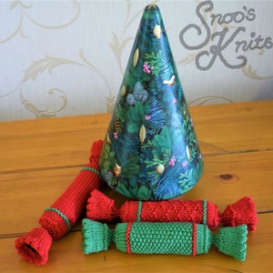 Puede incluir: Cuatro decoraciones de crackers navideños de punto. Los crackers son rojos y verdes con ribetes blancos. Los crackers están dispuestos en fila sobre una superficie de madera. Un adorno de árbol de Navidad verde y dorado está en el fondo.