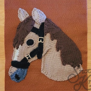 May include: A brown and white knitted horse head with a black bridle. The horse head is on a brown felt background.