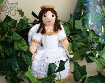 May include: A knitted angel doll with a white dress, gold trim, and brown hair. The angel is sitting in a bed of green leaves.