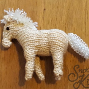 May include: A knitted beige horse with a white mane and tail. The horse is standing on a wooden surface.