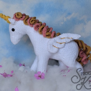 May include: White crocheted unicorn with pink and gold yarn mane and tail, and gold yarn wings. The unicorn is standing on a white fluffy surface with pink flowers.