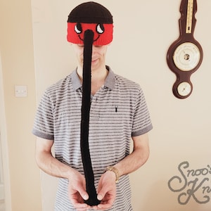 Op de afbeelding: Een persoon draagt ​​een rood-zwarte gebreide muts met een lange zwarte neus. De muts heeft een glimlachend gezicht met witte ogen en een zwarte neus. De persoon houdt de neus in zijn handen.