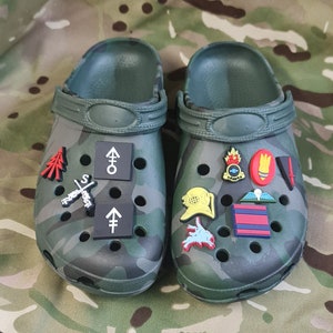 Puede incluir: Un par de zuecos de color verde con estampado de camuflaje con varios abalorios coloridos adjuntos. Los abalorios incluyen un árbol de Navidad rojo, una brújula blanca y negra, una cruz blanca y negra con un arma, una flecha blanca y negra, un submarino amarillo, una bandera roja y blanca, una bomba roja y blanca, una corona roja y blanca, una espada blanca y negra y un emblema de fuego rojo y blanco.