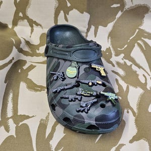 May include: A dark green camouflage clog shoe decorated with miniature weapon charms, including guns and a grenade. The shoe has a pivoting heel strap and is patterned in shades of green and grey.