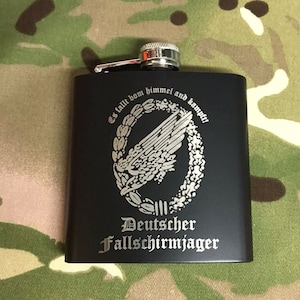 May include: Black metal flask with a silver cap and engraved text. The text reads "Es fällt vom himmel and kämpft" and "Deutscher Fallschirmjäger".