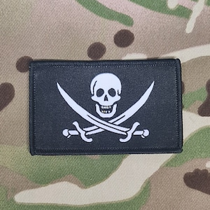 Könnte beinhalten: Ein schwarzer, rechteckiger Aufnäher mit einem weißen Totenkopf und gekreuzten Knochen, mit zwei gekreuzten Schwertern. Der Aufnäher befindet sich vor einem Tarnmuster-Hintergrund. Ein klassisches Piratensymbol.