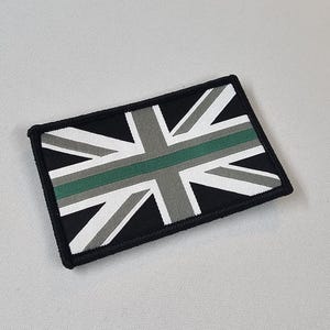 May include: A rectangular patch with a Union Jack design in black, white, grey and green. The patch has a black border. The design is a stylized representation of the British flag.