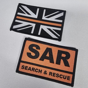 Op de afbeelding: Twee rechthoekige geborduurde patches. De bovenste patch heeft een zwarte achtergrond met een wit en oranje Union Jack-ontwerp. De onderste patch is oranje met de zwarte tekst "SAR" boven "SEARCH & RESCUE".