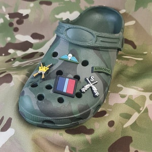 May include: A green camouflage clog with various military-themed charms, including a parachute, a rifle, and a flag. The clog has the word "AIRBORNE" printed on the side.
