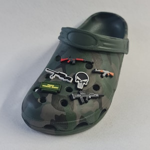 Könnte beinhalten: Ein grüner Camouflage-Croc-Schuh, verziert mit verschiedenen Charms zum Thema Waffen, darunter ein Totenkopf, eine Granate und der Text "FRONT TOWARD ENEMY". Der Schuh verfügt über einen schwenkbaren Fersenriemen und Belüftungslöcher.