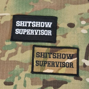 Può includere: Due toppe ricamate in bianco e nero con il testo "SHITSHOW SUPERVISOR" su uno sfondo mimetico.