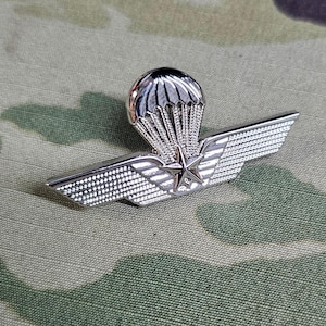 May include: A silver-toned military pin with a parachute and wings. The parachute has a star in the center.