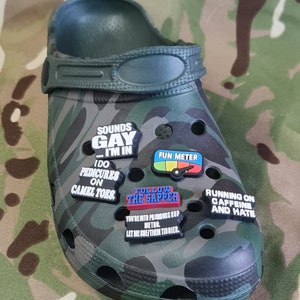 Può includere: Crocs di camuffamento verde con vari ciondoli per scarpe colorati. I ciondoli hanno frasi come "Sounds Gay...I'm In", "I Do Pedicures on Camel Toes", "Follow the Sapper", "Fun Meter" e "Running on Caffeine and Hate".