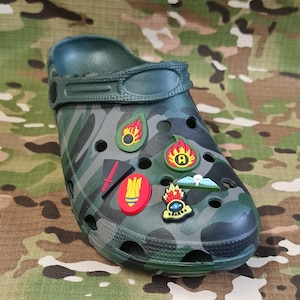 May include: Green camouflage patterned clog with various colourful cartoon character and symbol charms attached. The charms include a bomb, a sword, a flame, a star, a letter A, and a creature with an eye.