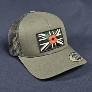 May include: An olive green trucker cap with a black mesh back. The cap has a rectangular patch with a black and white design and a red poppy flower. A small circular tag is on the side.
