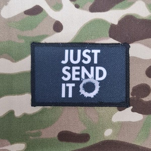 Può includere: Una toppa rettangolare con la scritta "JUST SEND IT" in lettere bianche su sfondo blu scuro. La toppa ha un bordo nero ed è posta su un motivo mimetico.