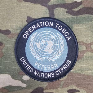 Puede incluir: Un parche circular con el texto "OPERATION TOSCA" y "UNITED NATIONS CYPRUS" en letras blancas sobre un borde negro. El centro presenta un fondo azul claro con un globo terráqueo y ramas de olivo. La palabra "VETERAN" también es visible.