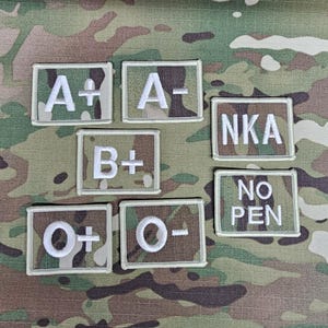 Puede incluir: Un conjunto de parches bordados con texto blanco sobre un fondo de camuflaje. Los parches muestran grupos sanguíneos como A+, A-, B+, O+, O-, y también incluyen "NKA" y "NO PEN".
