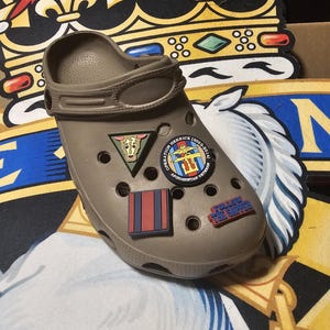 Peut inclure: Un sabot de couleur taupe orné de breloques décoratives. Les breloques comprennent un triangle avec une tête de taureau, un emblème circulaire avec du texte, un motif rectangulaire à rayures rouges et bleues et une breloque avec le texte "FOLLOW THE SIPPER".