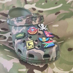May include: Clogs with a camouflage pattern and various colourful charms attached, including a skull and crossbones, a flag, and a military patch that reads "19th Airborne Market Garden".