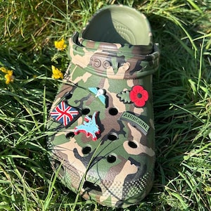 May include: Camouflage green Crocs with various charms, including a red poppy, a Union Jack flag, a blue bird, and a green 'AIRBORNE' charm.