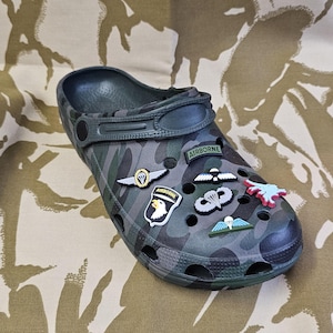 Puede incluir: Un par de zuecos de camuflaje verde con varios amuletos de temática militar, incluyendo un paracaídas, un casco alado y la palabra "Airborne".