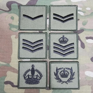 Puede incluir: Seis parches de rango militar sobre un fondo de camuflaje. Cada parche cuadrado tiene una base verde claro con insignias bordadas en azul oscuro. Los diseños presentan galones y coronas, que denotan varios rangos. Los parches están dispuestos en dos filas de tres.