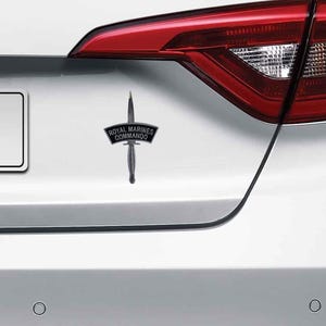 May include: A black and silver Royal Marines Commando car badge. The badge features a dagger design with the words "Royal Marines Commando" on a banner. The badge is attached to a white car.