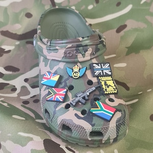 May include: A green and brown camouflage clog shoe decorated with several charms. The charms include flags, a small rifle, and a badge with wings and the letters "AD". The shoe is displayed on a camouflage background.
