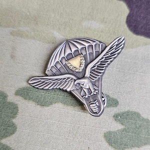 May include: A silver metal pin with a detailed design of a parachute and an eagle. The parachute is open and the eagle is flying out of it. The pin has a gold-colored accent on the parachute.