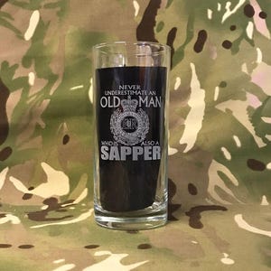 May include: A clear glass with a black background and white text that reads "Never underestimate an old man who is also a sapper". The glass features a white emblem with a crown and the letters "ER".