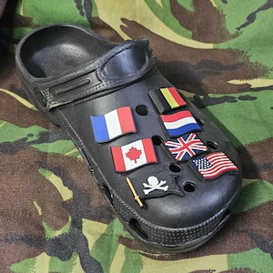 Könnte beinhalten: Schwarzer Clog-Schuh, verziert mit Flaggen-Anhängern, die Frankreich, Kanada, das Vereinigte Königreich, die Vereinigten Staaten, Belgien und die Niederlande repräsentieren. Ein Piratenflaggen-Anhänger ist ebenfalls vorhanden. Der Schuh befindet sich vor einem Camouflage-Hintergrund.