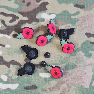 May include: A set of eight red poppy pins with green leaves. The pins are black and have a safety pin closure. The pins are on a camouflage fabric background.
