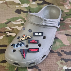May include: A pair of grey clogs with various military themed shoe charms. The charms include "Royal Navy Commando", "Army Commando", "Royal Marines Commando", "Ranger", and a sword.