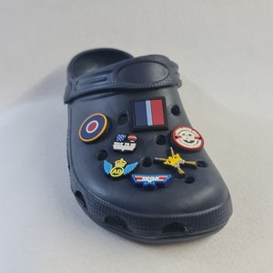 May include: A navy blue pair of clogs with various colorful charms attached. The charms include a red, white, and blue flag, a red circle with a white ring, a yellow and blue airplane wing with a crown and the letters "AD", a yellow and blue airplane with the text "Top Gun", a blue and white charm with the text "Talk to Me Wrangler", a yellow and red charm with the text "Maverick", and a yellow and red charm with the text "Danger Zone".