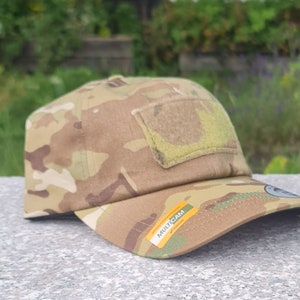 Może przedstawiać: Czapka bejsbolowa w kamuflażu z naszywką na rzep z przodu i żółtą etykietą z napisem "MULTICAM" z boku.