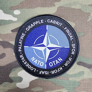 Op de afbeelding: Een ronde patch met het NATO-symbool in blauw en wit, omringd door tekst waaronder "NATO" en "OTAN". De patch heeft een zwarte rand met witte letters, op een camouflage achtergrond.
