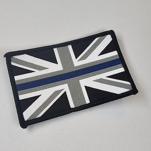 Può includere: Toppa rettangolare con un design Union Jack stilizzato. La bandiera incorpora elementi bianchi, grigi e neri, con una striscia blu al centro. Bordo nero.