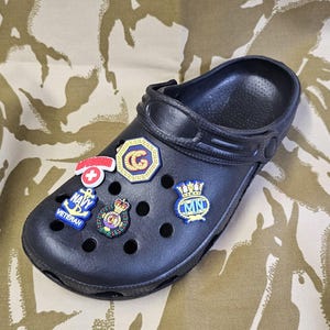 May include: Black clog with various colorful charms attached. The charms include a red cross, a gold crown, a blue anchor with the word "NAVY VETERAN", a gold crown with a red and gold design, and a blue and gold charm with the letters "MN".