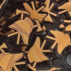 Op de afbeelding: Een verzameling zwarte en bruine schoenbedels. De bedels hebben ontwerpen met een brandweerthema, waaronder helmen, bijlen en andere symbolen. De bedels zijn gemaakt van een rubberachtig materiaal en zijn ontworpen om aan schoenen te worden bevestigd.