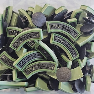 May include: A collection of olive green and black RAF Regiment badges. The badges are curved with the text "RAF REGIMENT" in a white and gray font. The badges are mixed with black, circular fasteners.