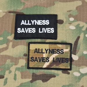 Può includere: Due toppe ricamate in bianco e nero con il testo "ALLYNESS SAVES LIVES" su uno sfondo mimetico.