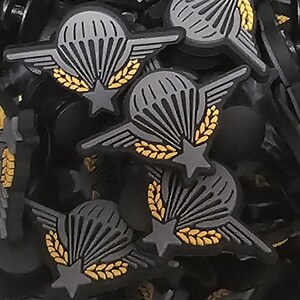 Pode incluir: Uma coleção de amuletos pretos e cinzentos com tema de paraquedas. Cada amuleto apresenta um design de paraquedas cinzento com uma estrela, asas e folhas de louro douradas. Os amuletos são provavelmente para artesanato ou fabricação de joias.