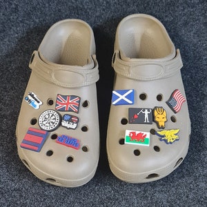 Peut inclure: Une paire de sabots beiges avec divers charmes de chaussures colorés. Les charmes comprennent des drapeaux, une boussole, un crâne et des os croisés, et le texte "Follow the Sapper".